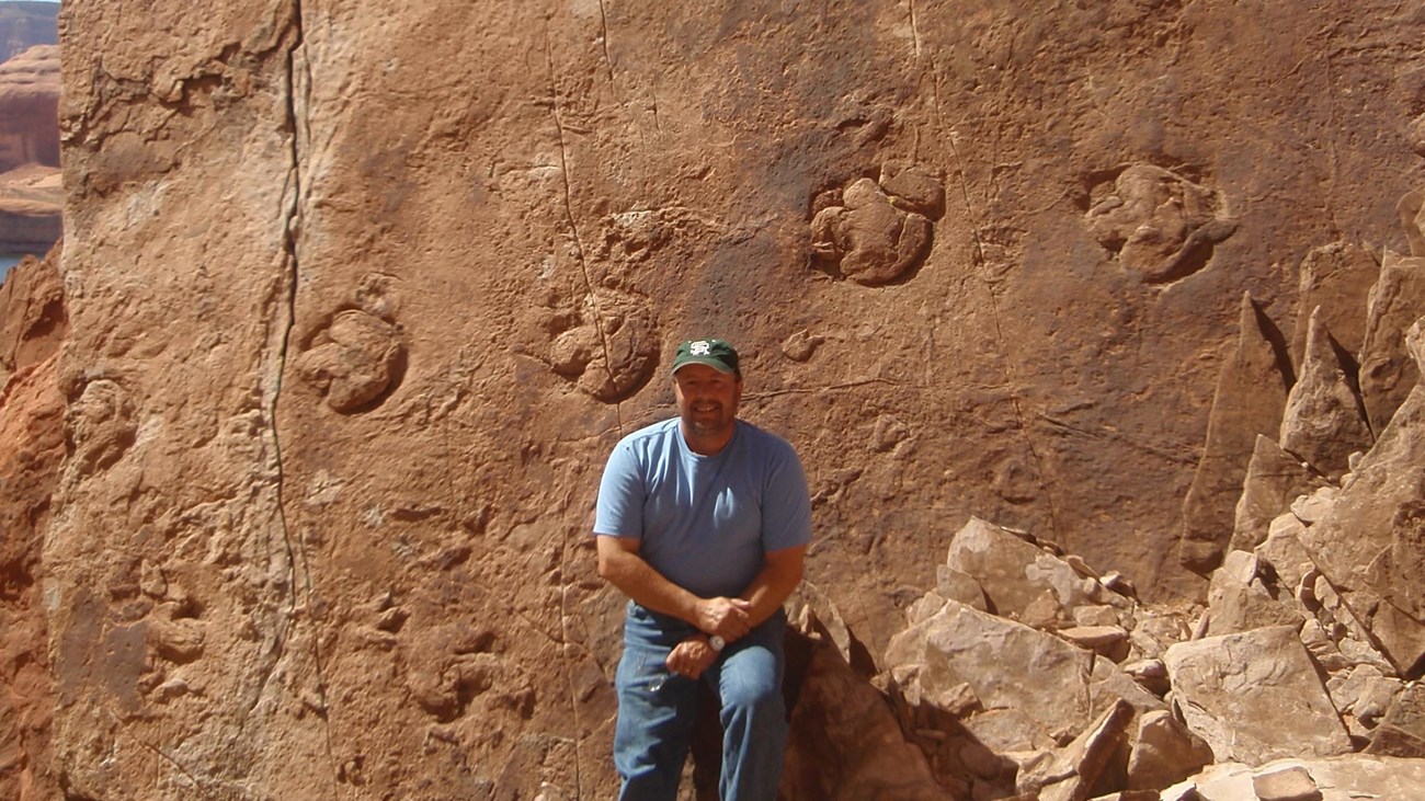 Photo of a person sitting in front of a large boulder slab featuring fossil dinosaur tracks.