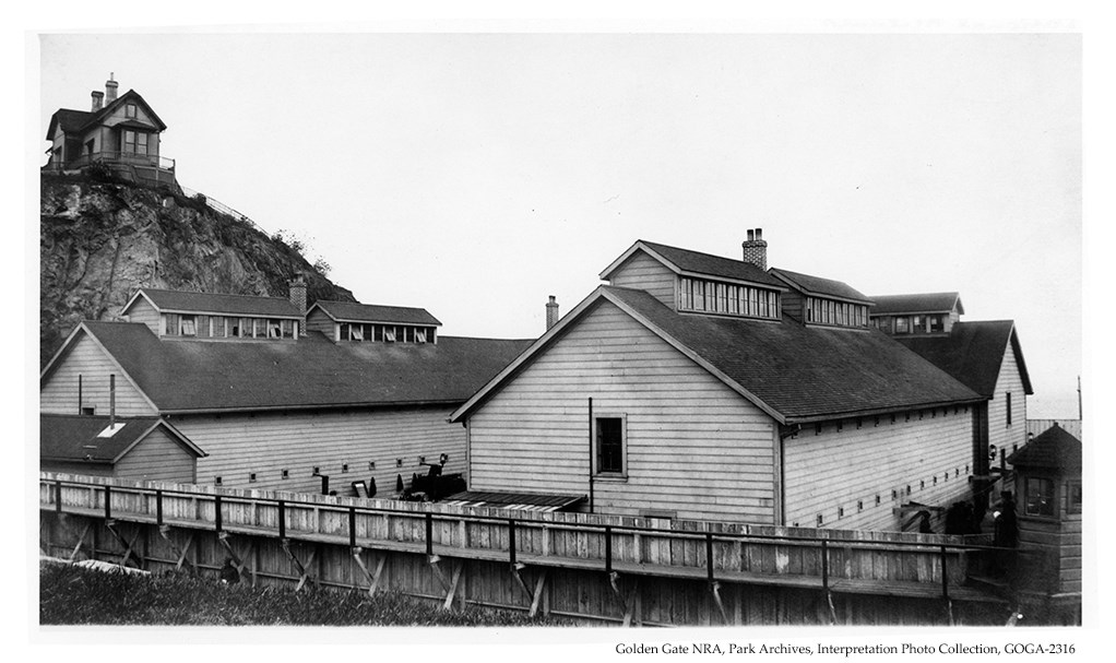 Black and White image of buildings surrounded by a wooden fence and guard tower. A house sits on top of a hill in the background.