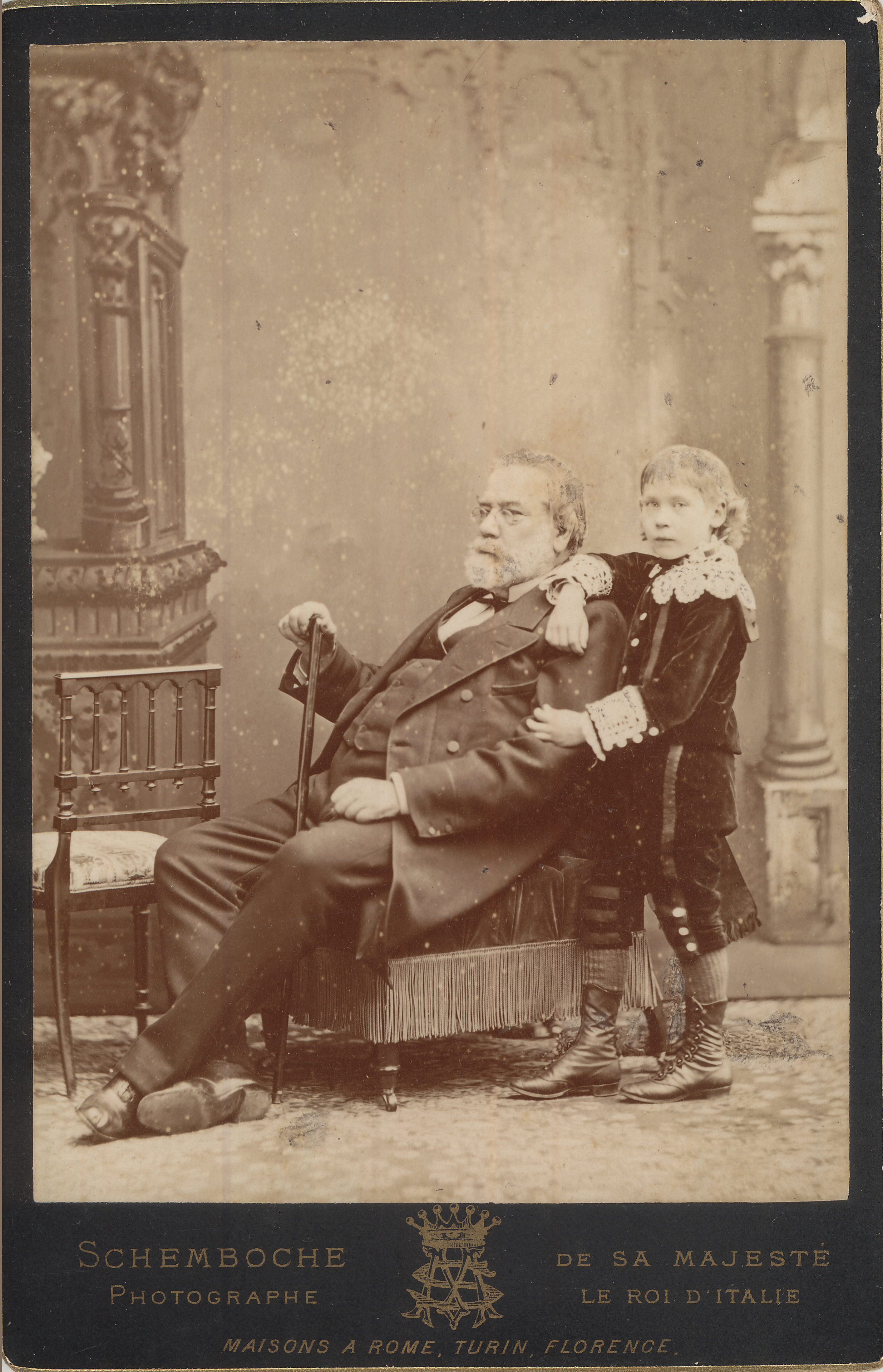 historic black and white photo of older man sitting in a chair and a young boy standing behind him, text at bottom of photo reads "Schemborche Photographe, De Sa Majeste Le Roi D'Italie, Maisons A Rome, Turin, Florence"