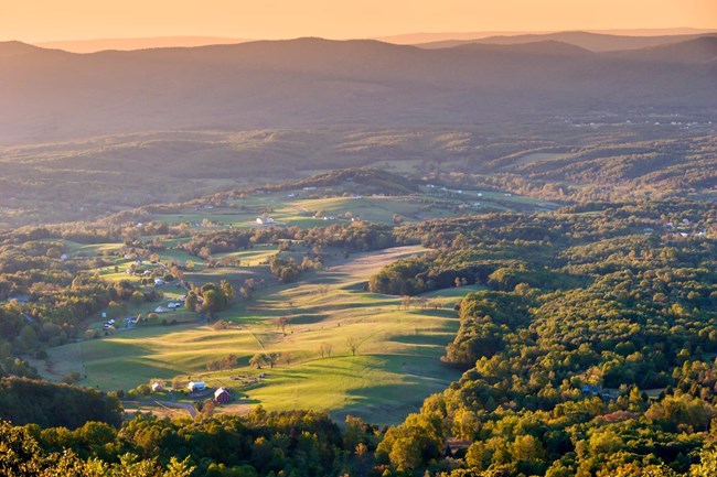 An aerial view of a valley landscape dotted by large fields, pastures, knots of trees, and clapboard farmhouses. Densley forested, rolling hills and mountain peaks can be seen in the background.