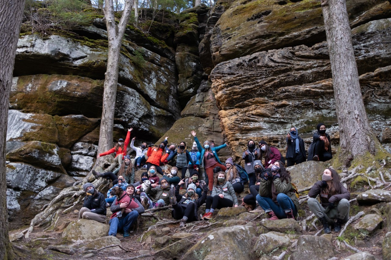 Two dozen women in hiking clothes and two dogs pose at the base of a sandstone cliff.