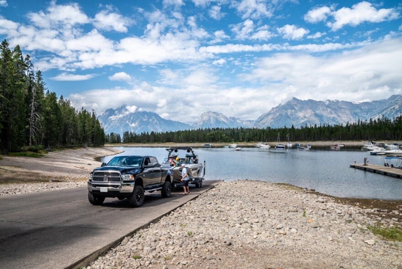 A truck backs a trailer with a boat down a boat ramp on a reservoir with mountains in the background