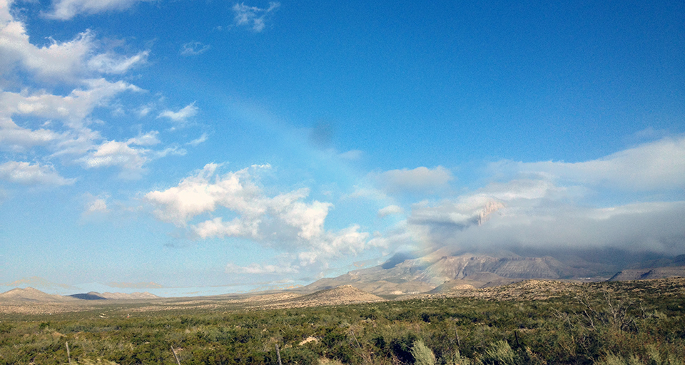 A rainbow over a shrubby desert landscape with cloud-covered, rugged mountains in the background.