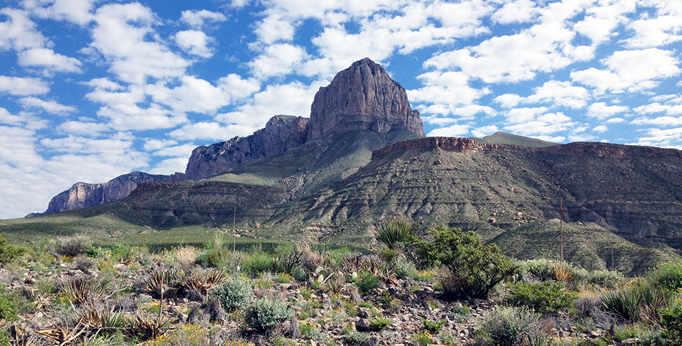Mountain peaks of sheer rock faces above a shrubby desert floor under scattered, cottony clouds.
