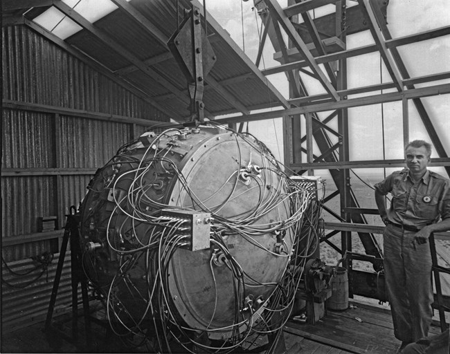 Black and white photo of a bomb covered in wires. A man in coveralls leans in the right of the photo. A