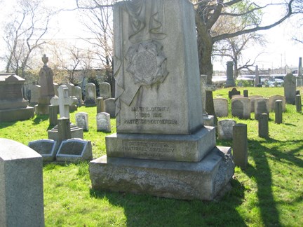 Large, light grey monument, in a cemetery, surrounded by gravestones