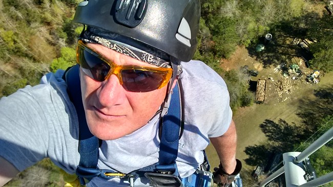 A man wearing a helmet, sunglasses, and climbing gear takes a selfie from the top of a 300 foot tower.