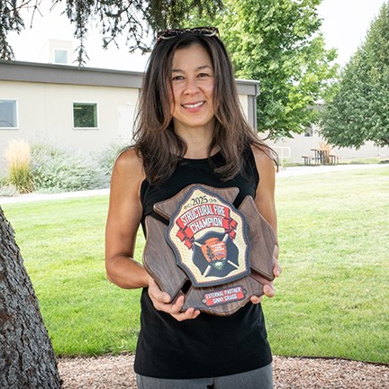 Ginny Gragg stands beneath a coniferous tree holding a plaque.