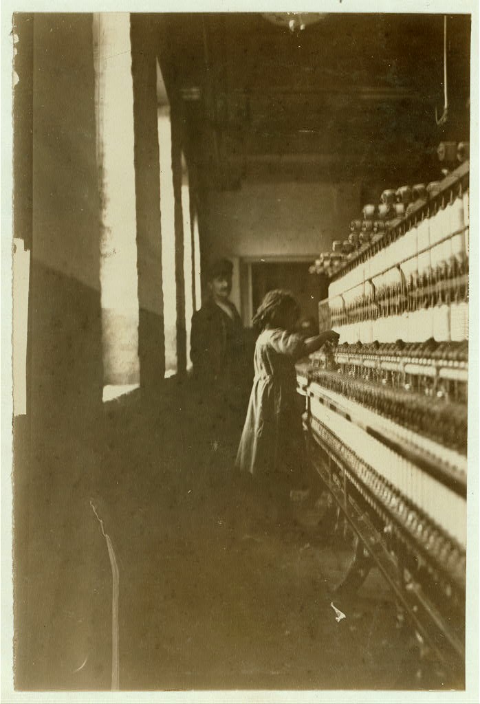 Yellowed photo of a young girl at work at a textile mill machine as man in a suit and hat looks on.