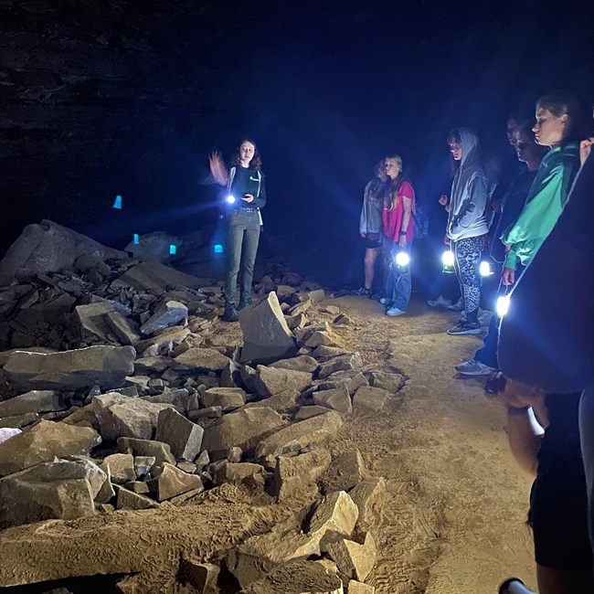 A person talks to a group of young women holding lanterns inside a rocky and dark cave.