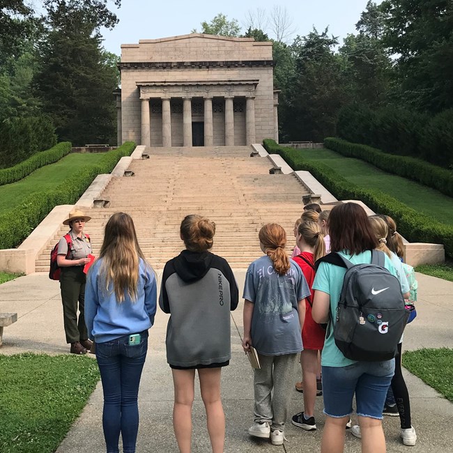 A group of people face a long and wide stairway that leads to a large stone building with large columns.