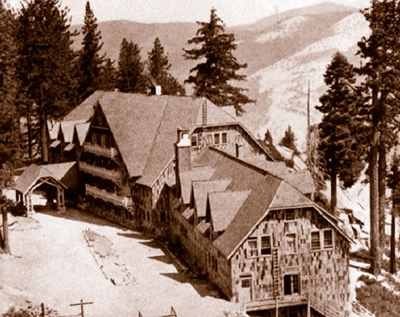 Sepia-tone photo of a large lodge with trees and mountains nearby.