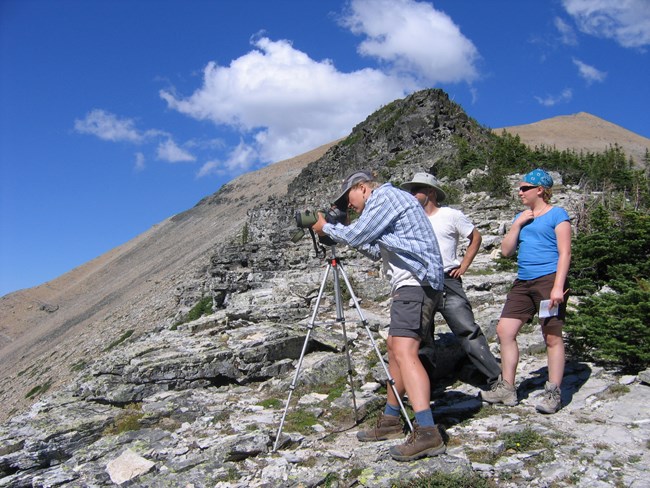 A group of three people stand below a mountain next to a scope with a tripod extended. One person looks through the scope and the others look into the distance.