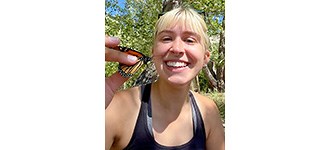 Kowalski in a woodland, holding up a monarch butterfly and smiling for the camera.