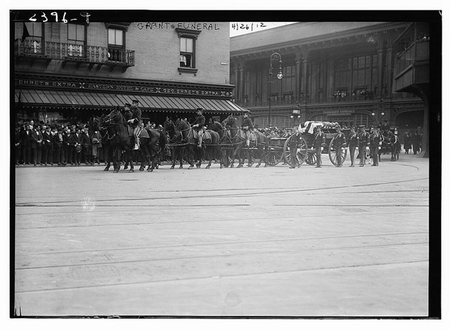 Black and white photo of horses with men on them pulling a dark carriage along a street with people in the background curb watching