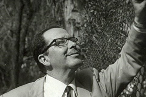 Black and white photo of a man in glasses and a suit smiling as he looks up leaning against a fence