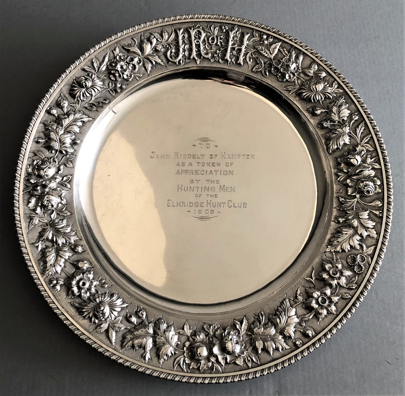 A circular silver tray with engraved art around the edges and an inscription in the middle.