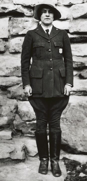 Polly Mead in the standard uniform stands with her hands down at either site and a wears a floppy hat.