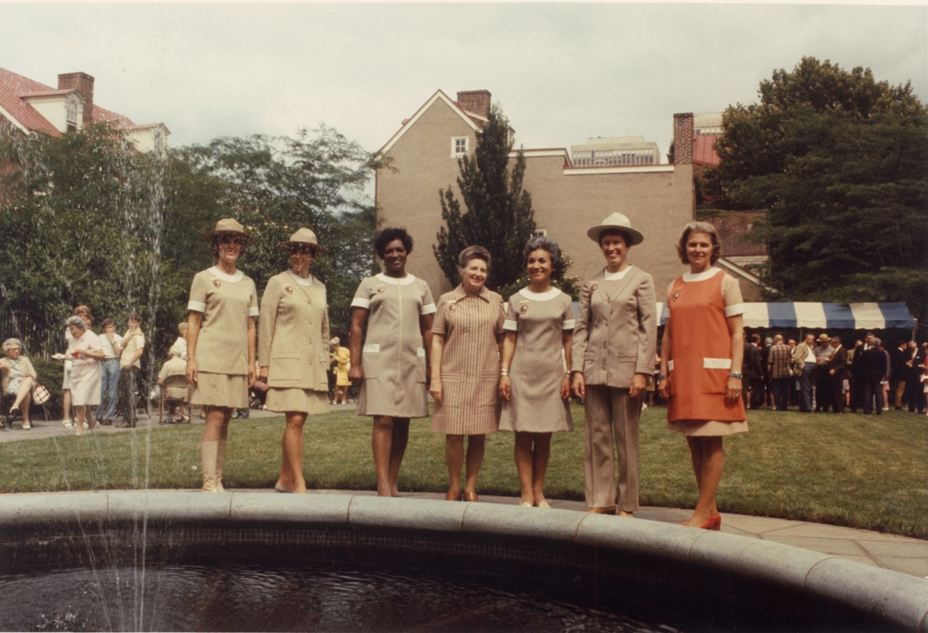 Seven women stand in a row modeling beige and orange uniforms