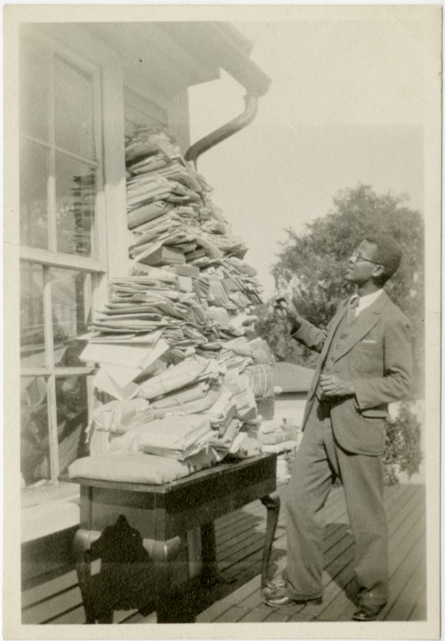 A Black man in a suit looks up at a pile of papers sitting on a desk that tower next to him.