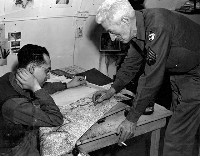 A seated man looking at a map while a white-haired older man points at the map.