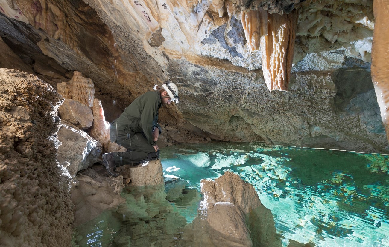 A uniformed National Park Service employee with a helmet and light kneels on a cave floor overlooking a cave pool