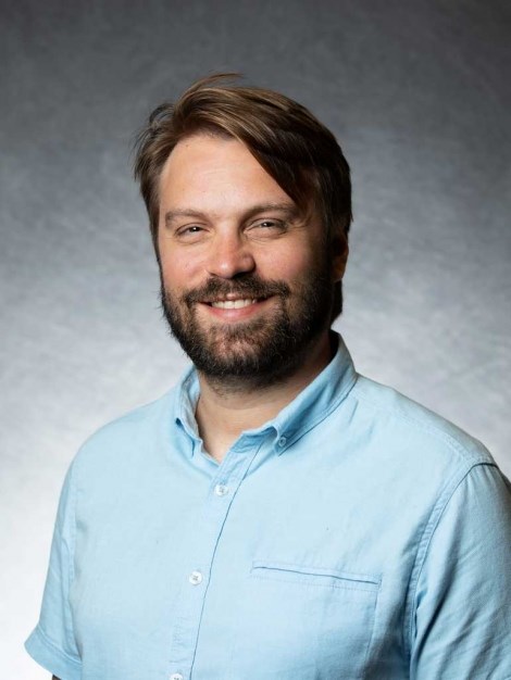 Headshot of a white man with brown hair and a beard wearing a blue shirt