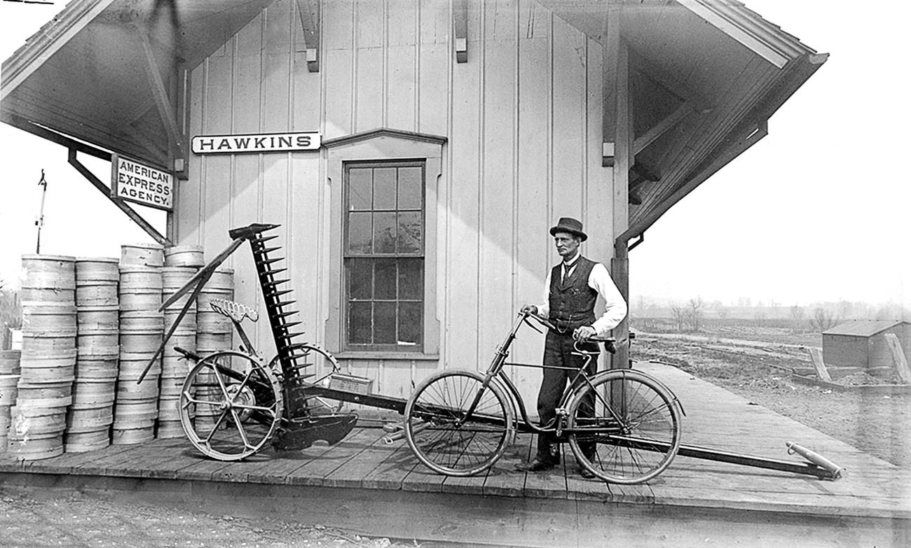 A man in a vest stands beside his bicycle on a railway platform labeled Hawkins; to the left are tall stacks of round wooden cheese crates.