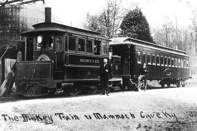 A black and white photo of two men standing on or near a small train engine with “Hercules” written on its side and passenger car. Text written along the photo states “The Dinkey Train to Mammoth Cave Ky”.
