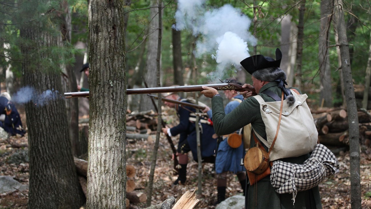 Park Service Volunteers are shown demonstrating Revolutionary War combat techniques in a wooded area.