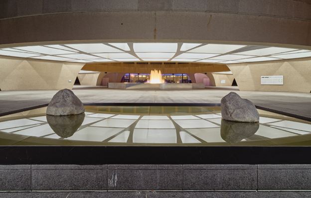 A wide horizontal open-air courtyard with a water fountain with stones and reflecting overhead light panels.