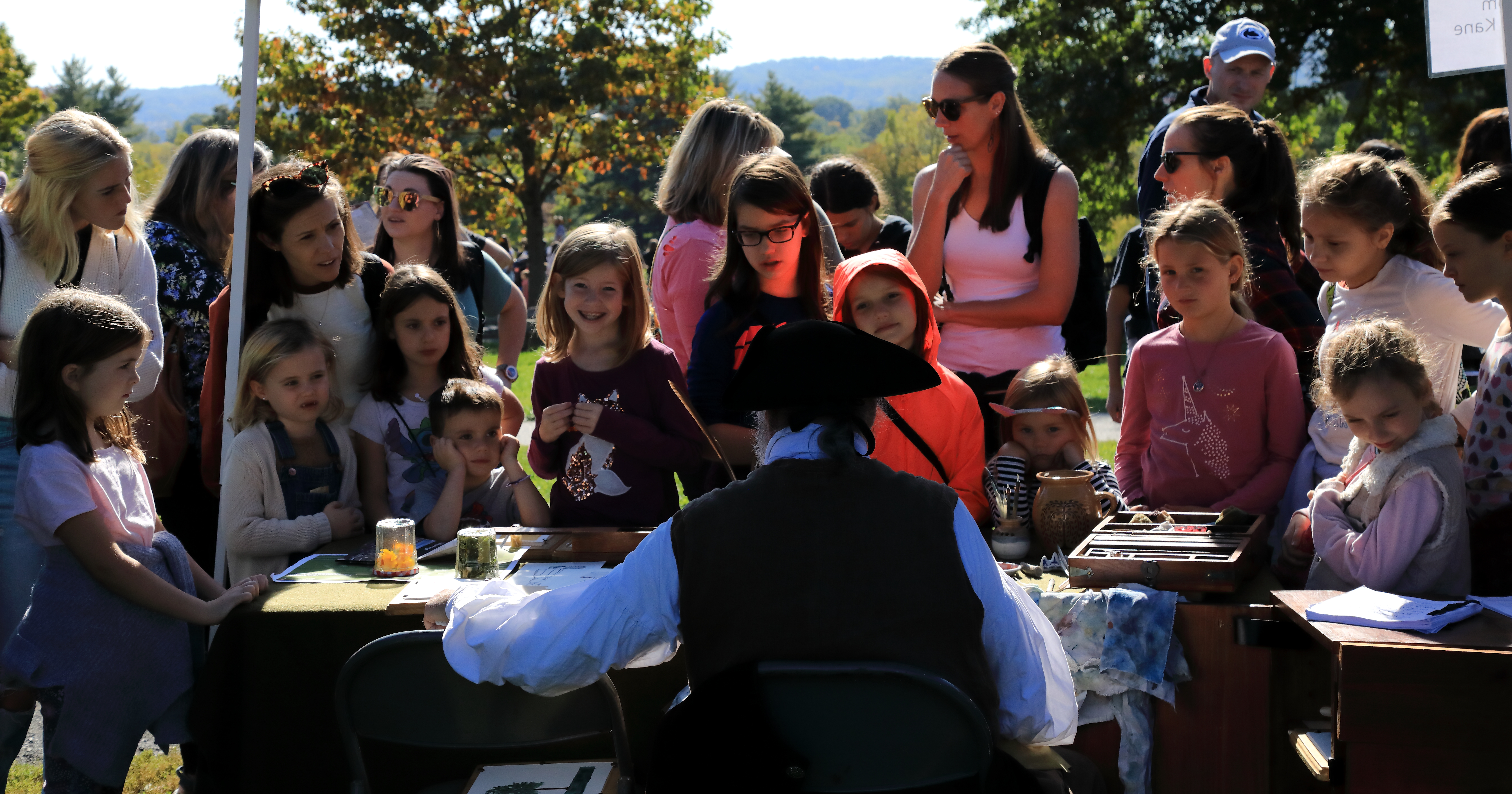 a large group of children gather in front of a table where a man wearing 18th century clothing speaks to them