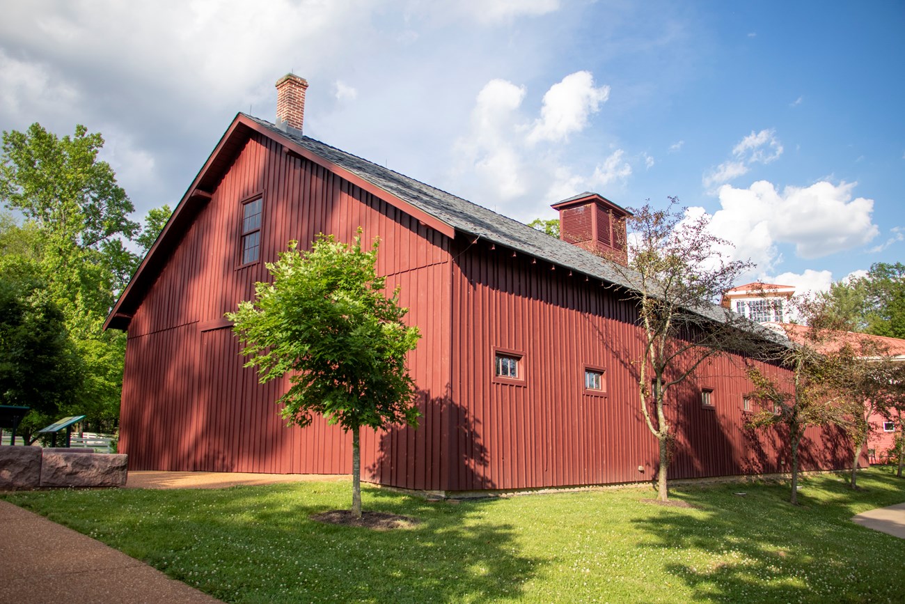 Red two-story frame stable made of wood. Four small trees and a walkway are in front of the horse stable.