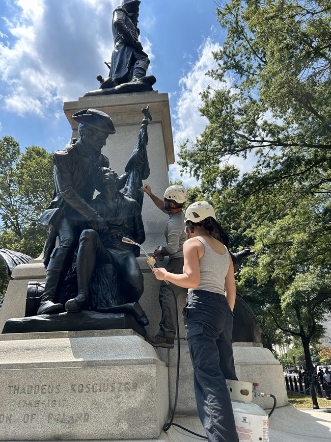 Woman holding hot waxing tool and man in background. They are working on the bronze statue of Brigadier General Thaddeus Kościuszko.