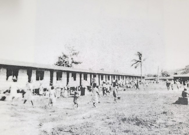 Black and white photo of long, single story housing with people in courtyard