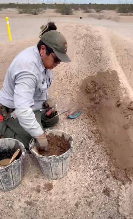 A person in uniform kneels on a gravel surface, using their hands to mix a brown adobe-like material in a metal bucket.