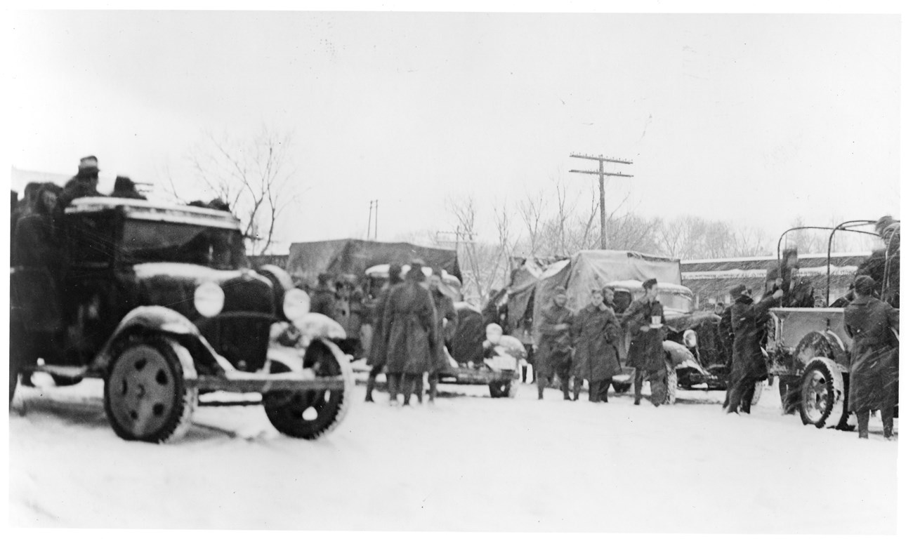 Uniformed men in long coats gather around four army trucks being unloaded on a snowy day.