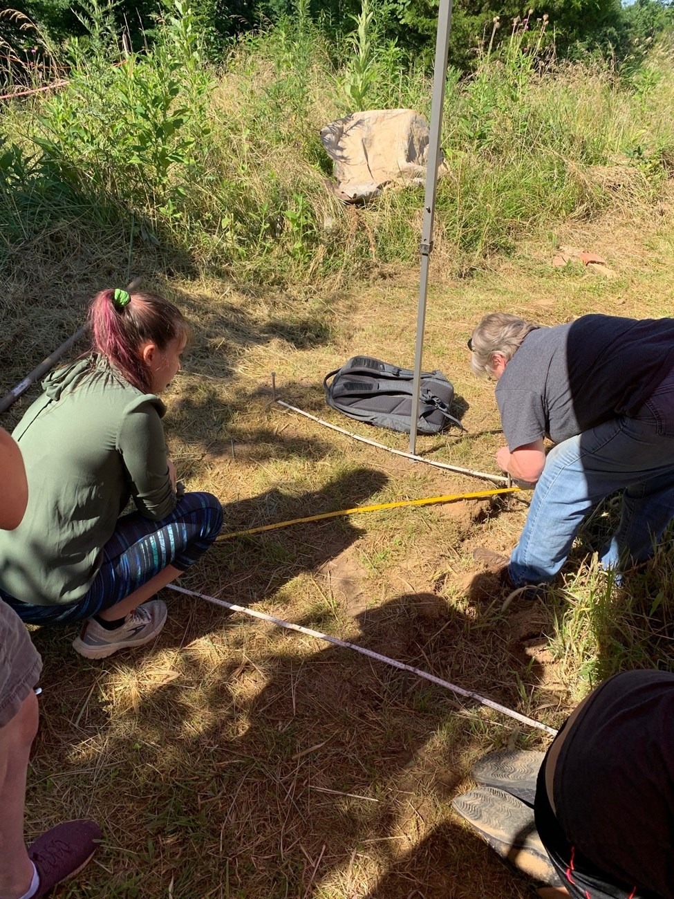 A high school student watches as an experienced archaeologist begins to dig in the ground.