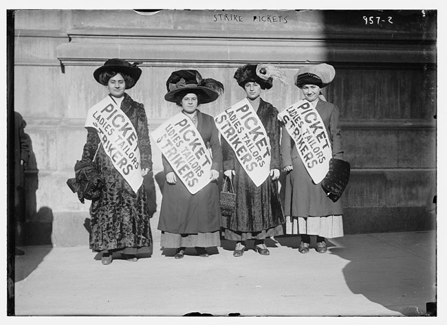 Four women in hats and dresses wearing banners that read "Picket Ladies Tailors Strikers"