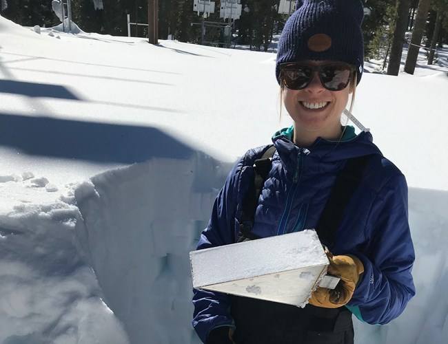 Woman standing in snow pit and showing a triangular metal tool used for sampling snow