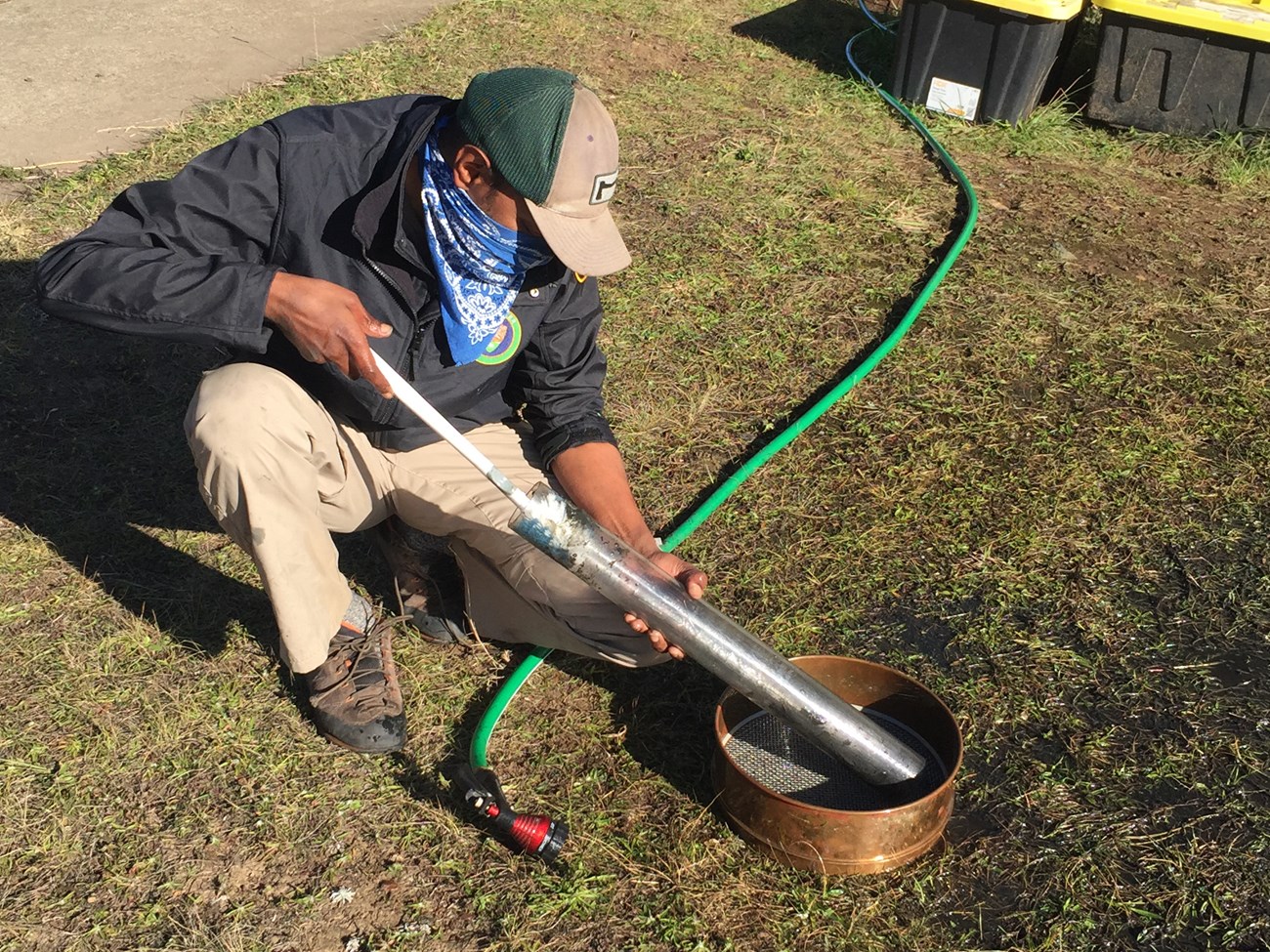 Person kneeling on the ground, holding a tube full of dark sediments over a mesh sieve.