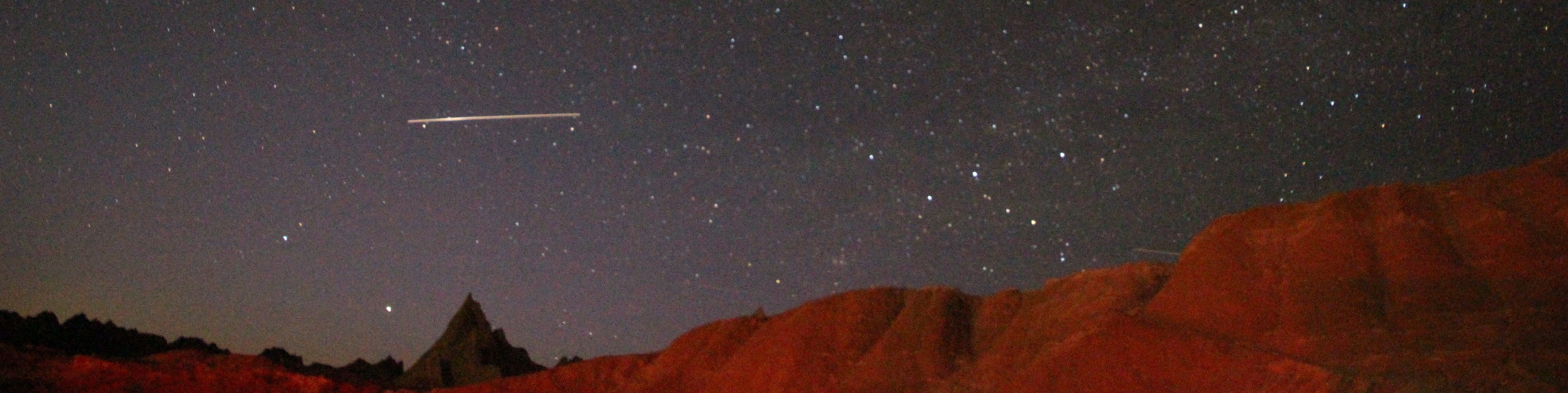 a starry night sky with a white streak in the left corner, appearing just over some red badlands buttes
