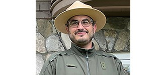 Malzone outside of a building in a National Park Service hat and jacket, looing past the camera with a smile.