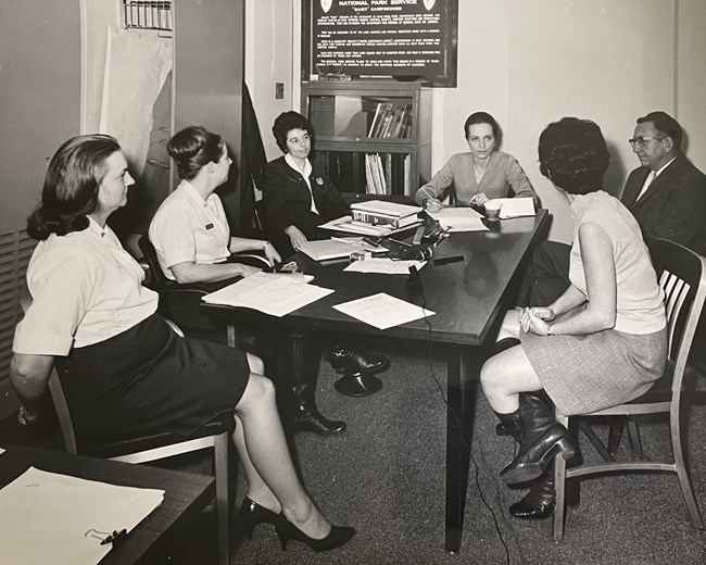 Half a dozen women sitting around a table, looking towards the woman at the the head of the table.