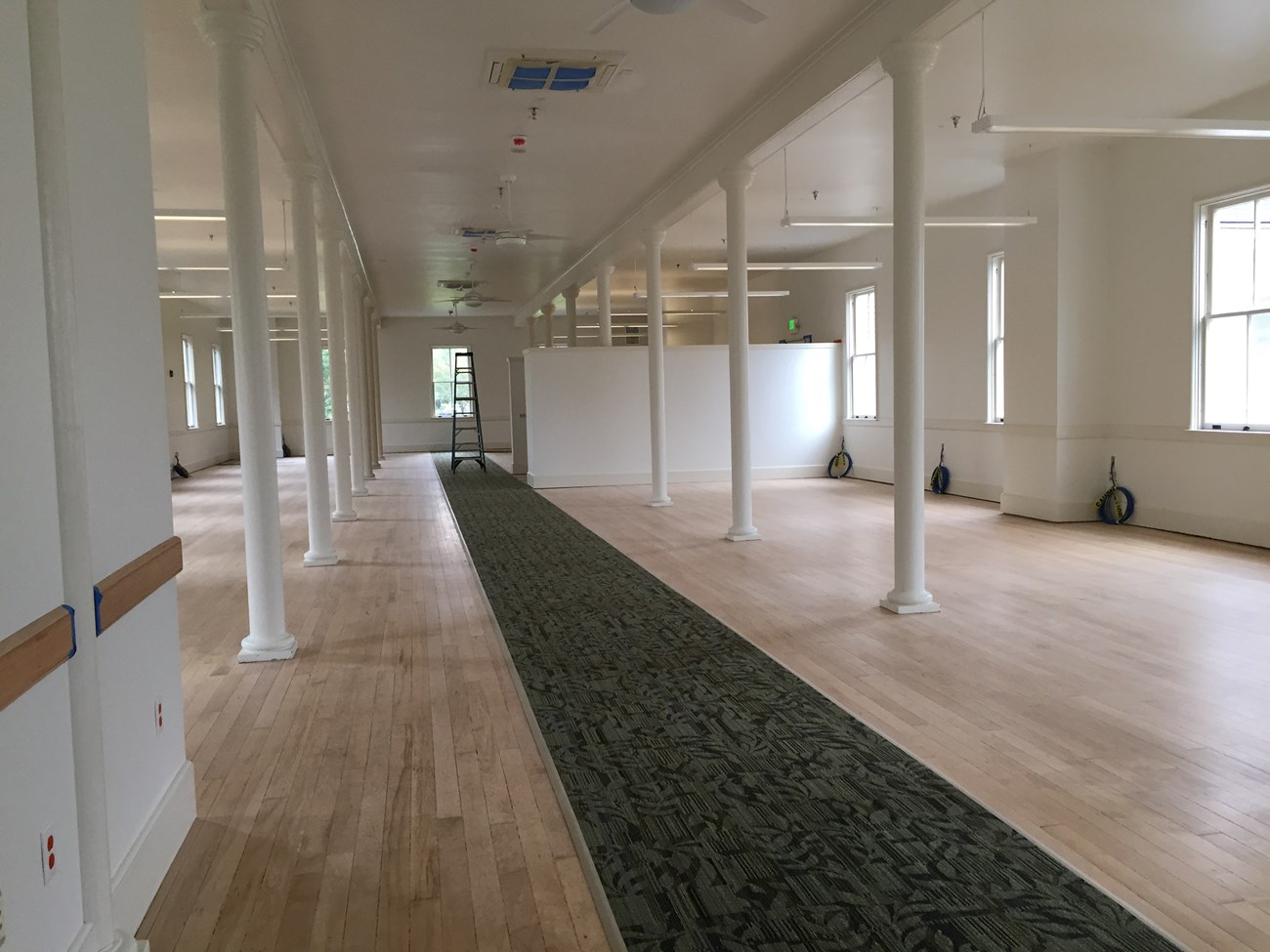 A large, open room with cream-colored walls and a wooden floor. The room is divided by two rows of thin white columns and has a dark-colored carpet runner down the middle. Many tall windows line the sides of the room.