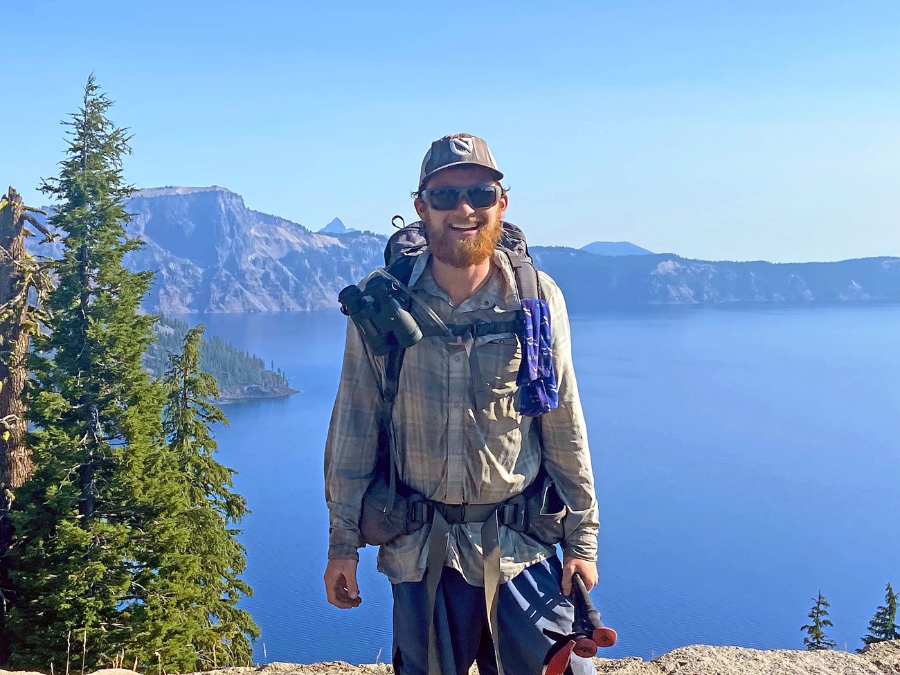 The author, with sunglasses and a beard, smiles for a photo in front of Crater Lake, which appears faded in haze from wildfire smoke. He carries a large backpacking pack, binoculars, and a pair of hicking poles.