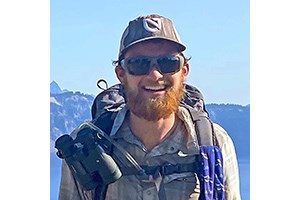 Headshot of will in hiking gear, wearing a baseball cap, backpack, sunglasses, and a beard.