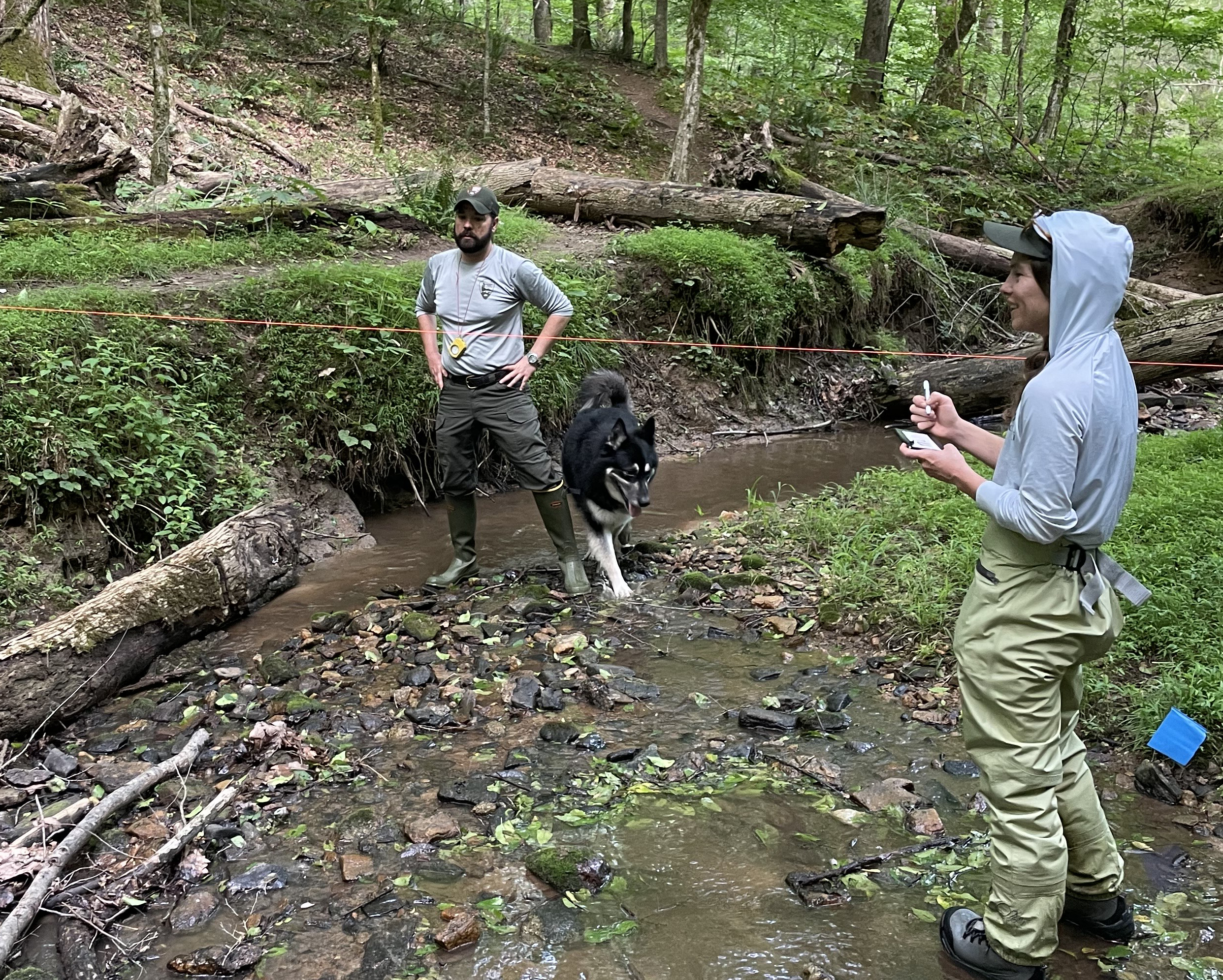 man holding a camera and woman holding a dry erase card standing in a stream with a black dog