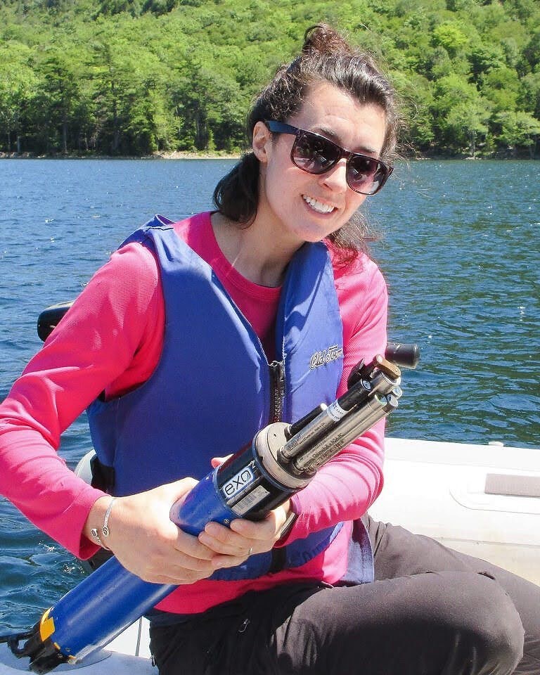 Person in a boat wearing sunglasses, a pink long-sleeved shirt, and a life jacket, holding a large, cylindrical device over the water.