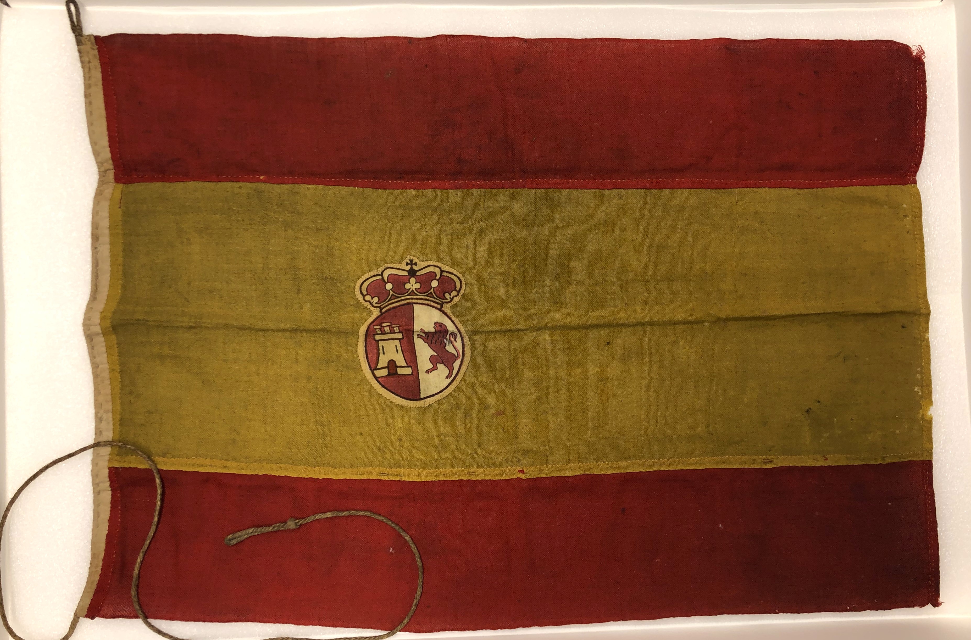 A photo of a Spanish flag, with two red stripes and one yellow stripe. The seal of Spain is located in the yellow stripe. The flag is worn.